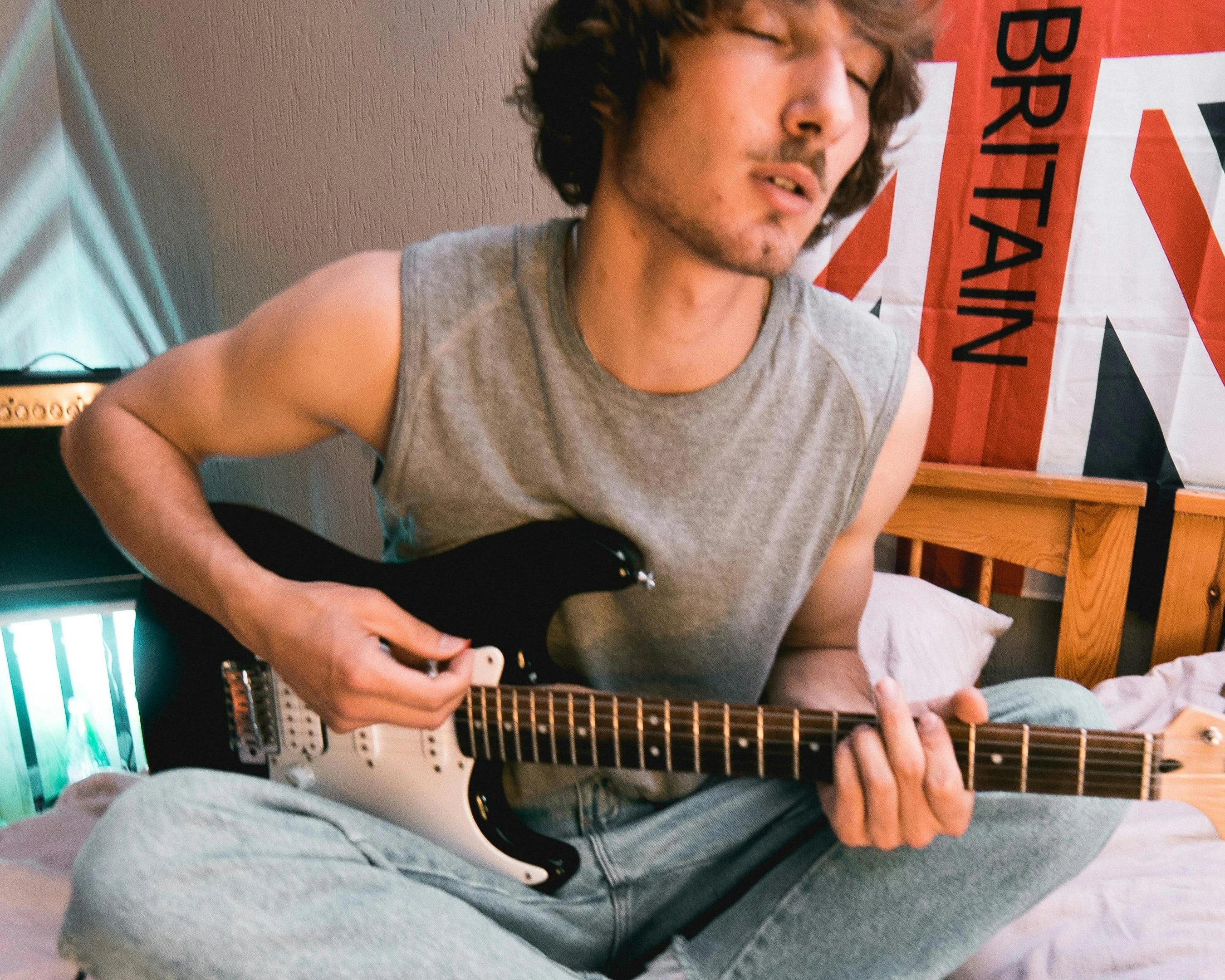Young man sitting cross-legged on bed playing Fender Stratocaster.