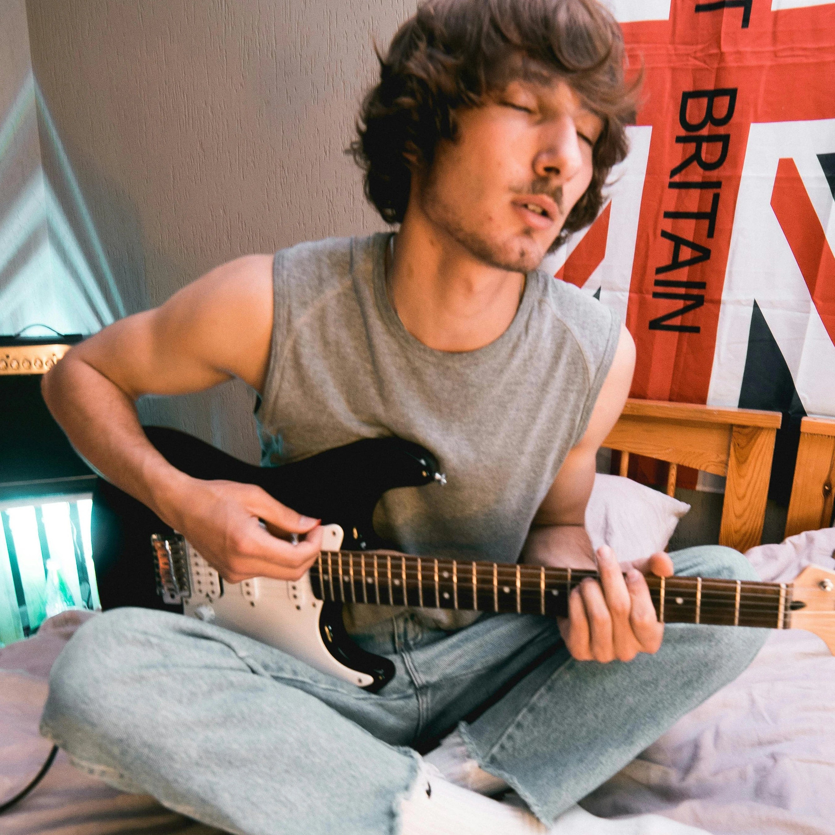 Young man sitting cross-legged on bed playing Fender Stratocaster.